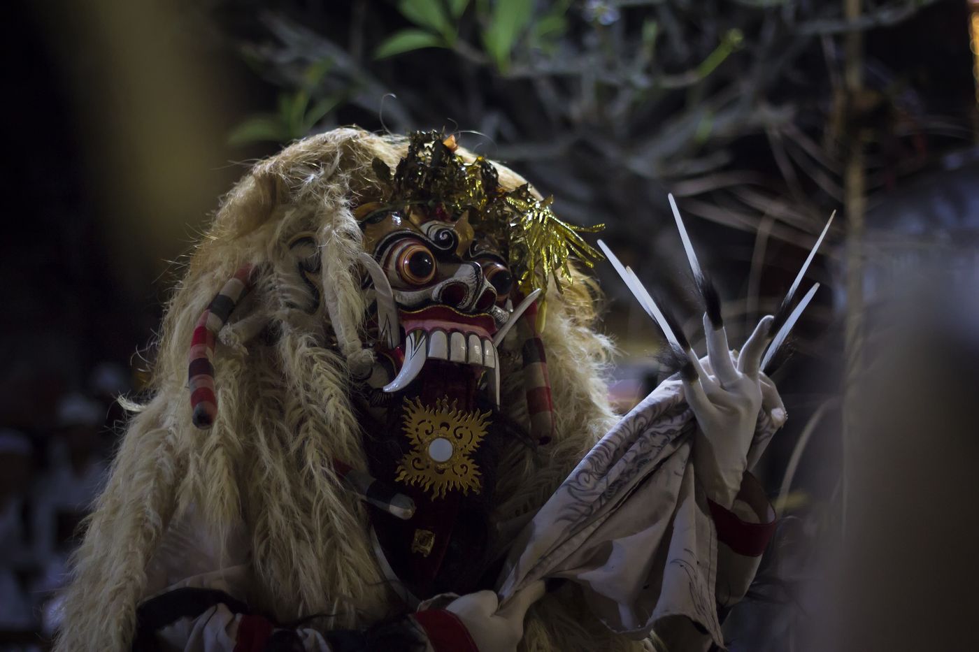 A dancer in the Rangda mask, with long pale hair, bulging eyes, tusks and clawed white hands, performing at night