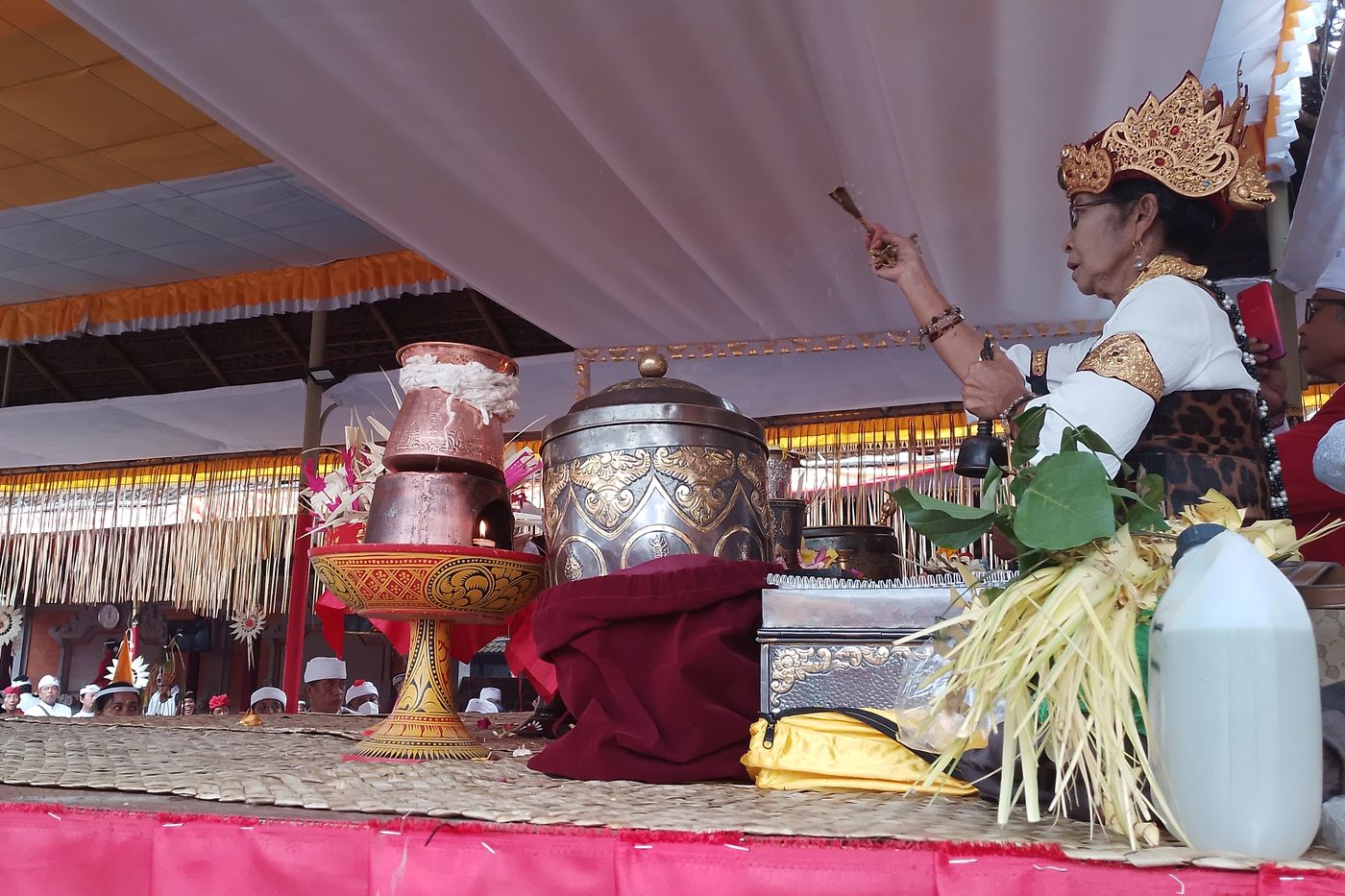 A woman high priest in a gold crown, a bell in one hand and a ritual implement raised in the other, behind holy-water vessels at a tawur ceremony