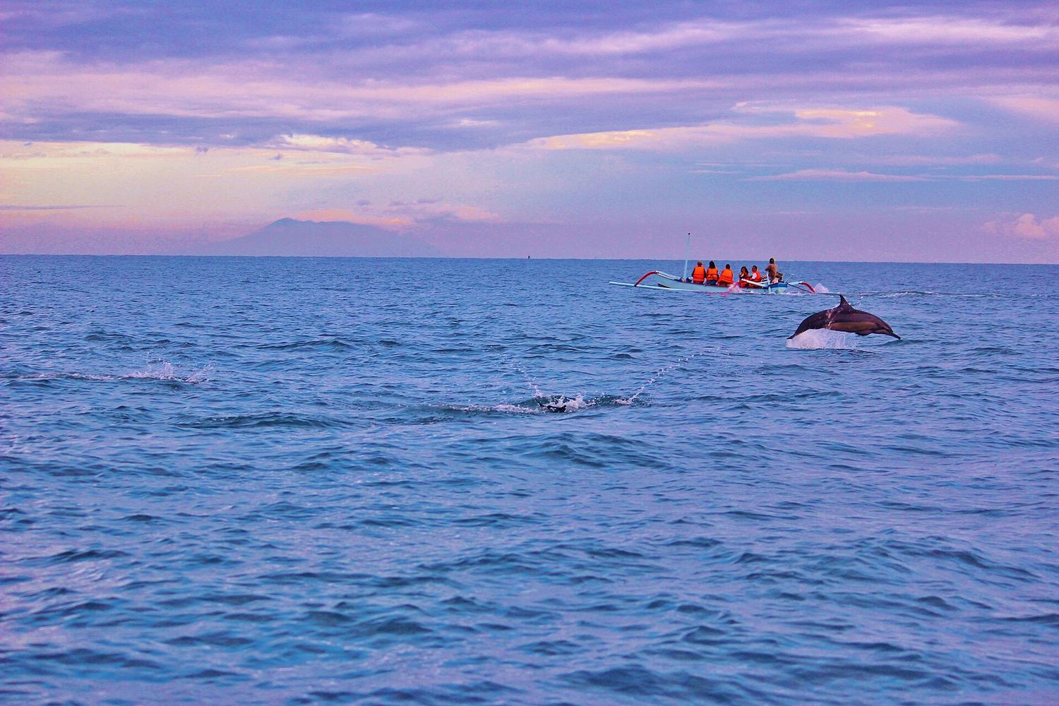 A dolphin leaping beside a jukung outrigger boat carrying passengers in orange life jackets, at sunrise off Lovina