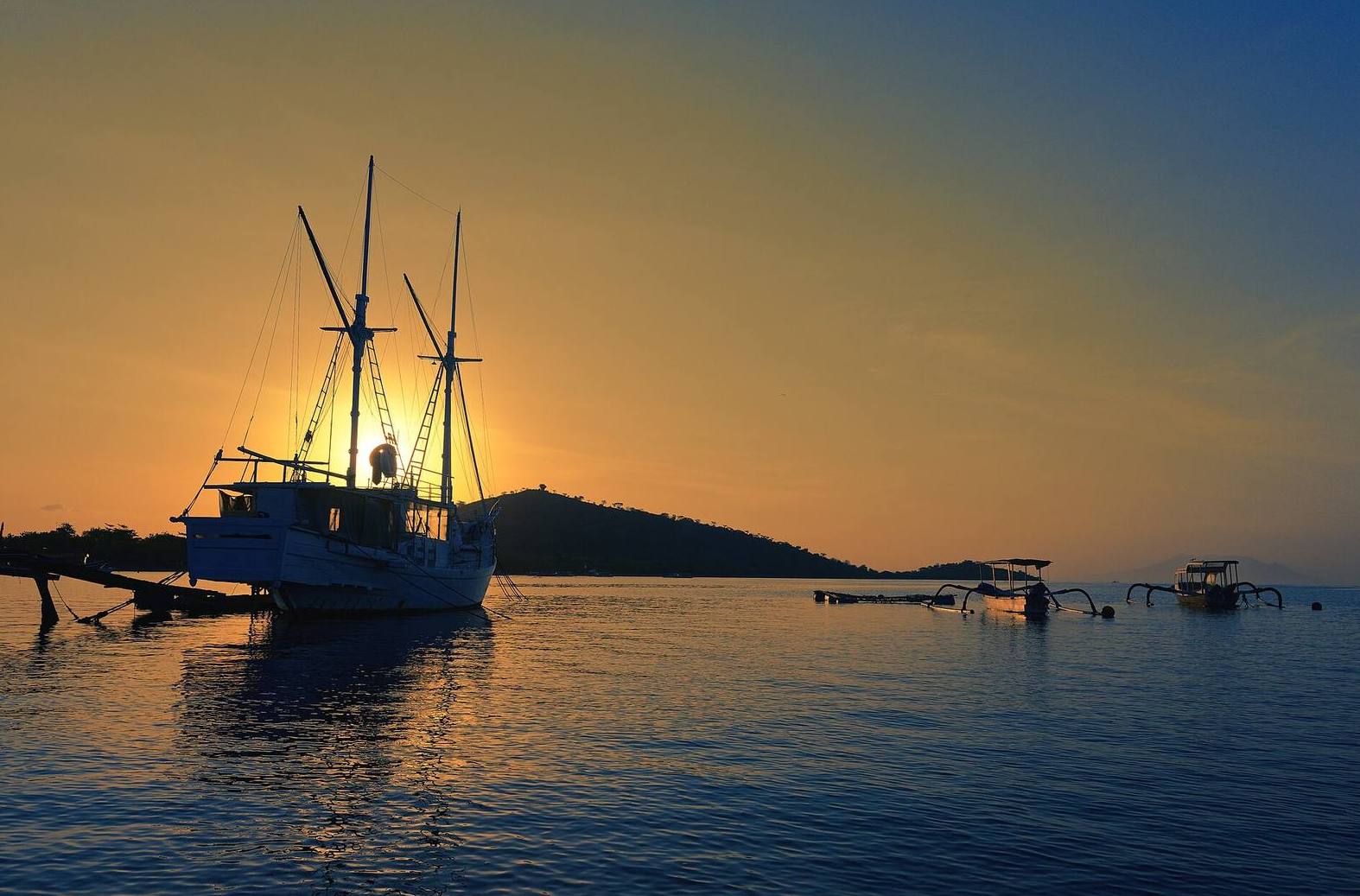Low sun over the calm bay at Pemuteran, a wooden boat and outrigger boats at anchor and a hill across the water